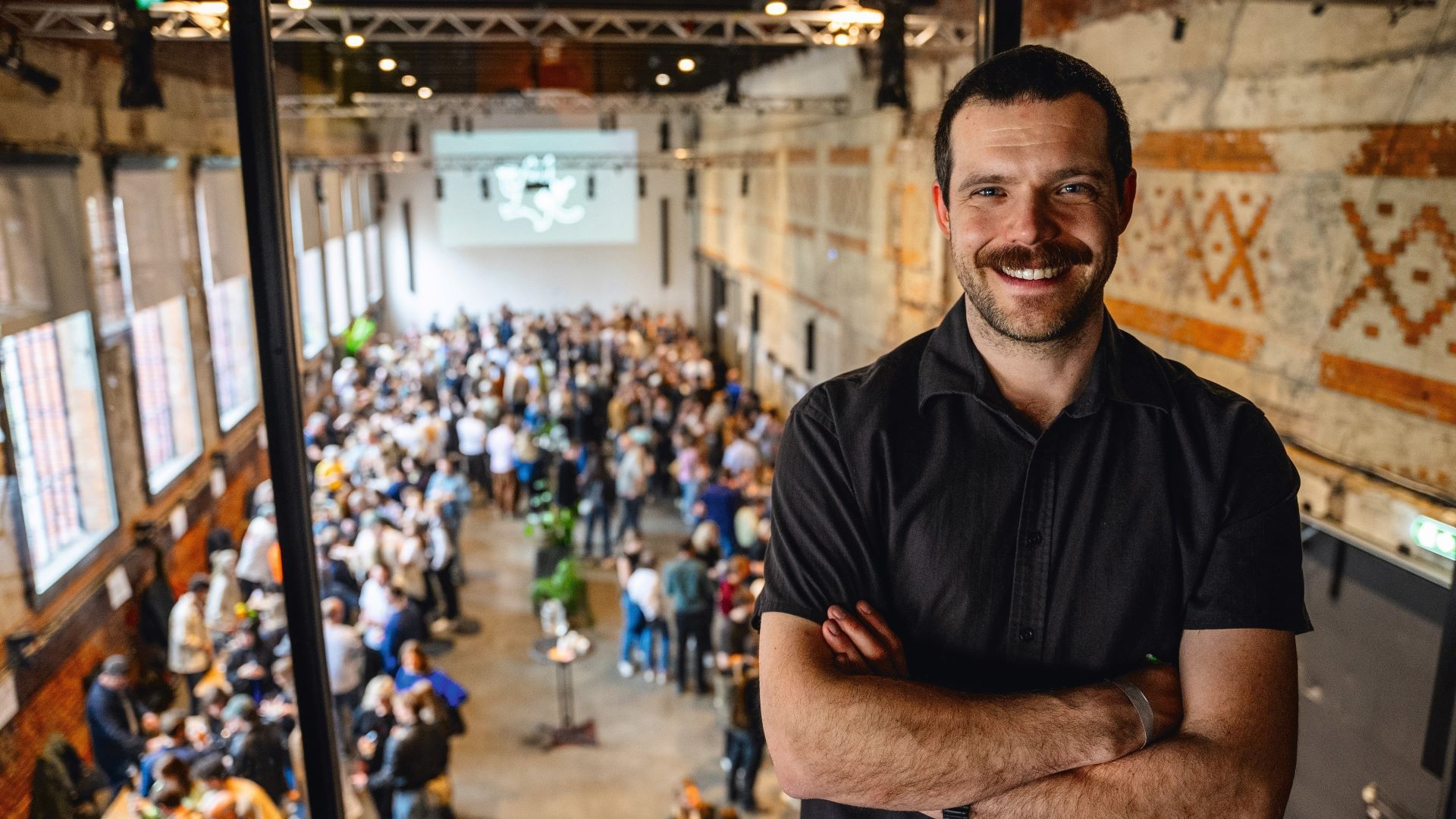 Mann som står med armene i kryss og smiler til kamera. I etasjen under han foregår en naturvinmesse med mange besøkende.