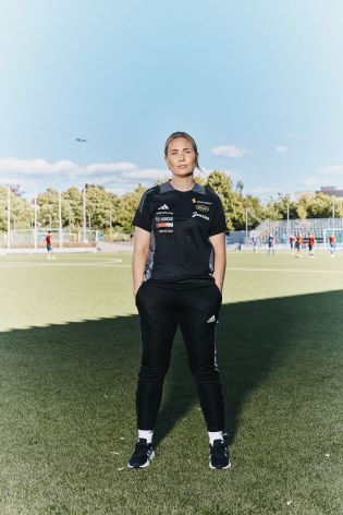 Kvinne som står på en fotballbane med hendene i lommene, mens det spilles en kamp bak henne.